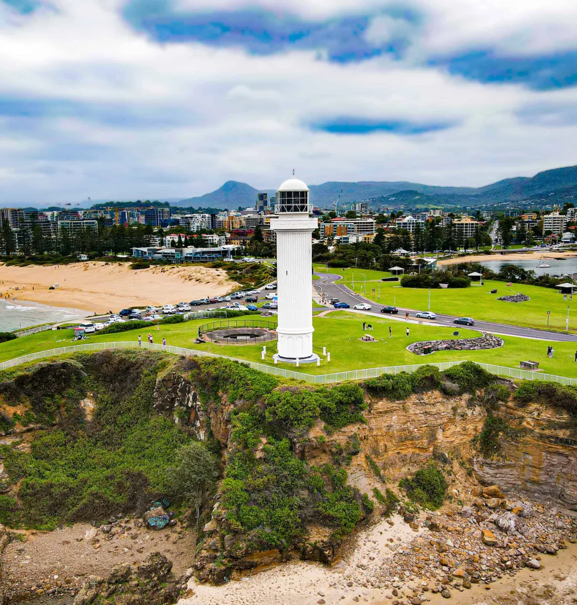 Lighthouse on a cliff overlooking the city and beach, with green park and cloudy sky.